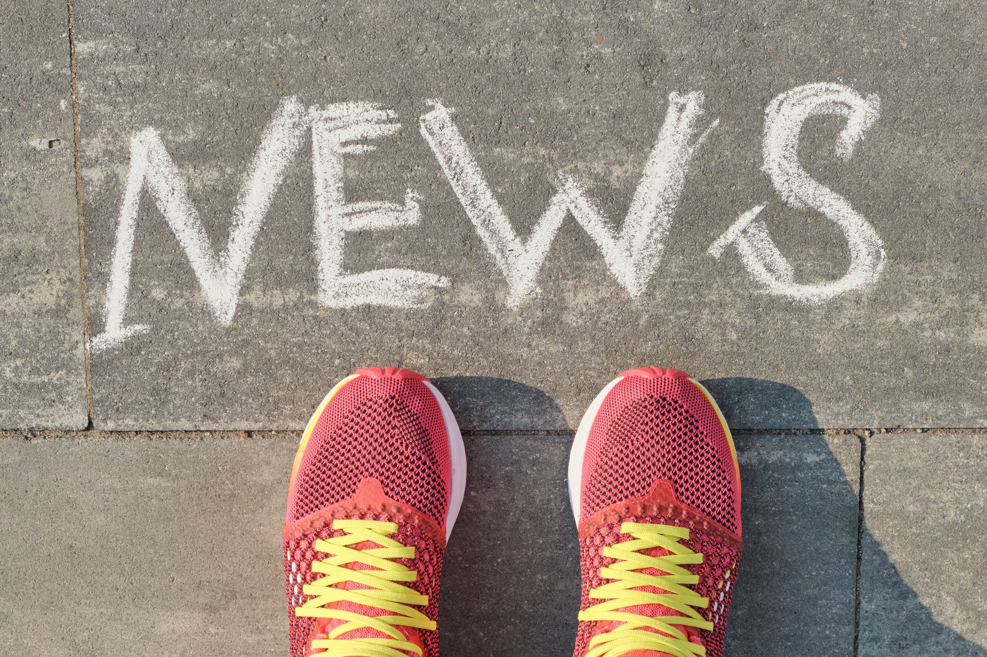 Word news on gray sidewalk with women legs in sneakers, top view